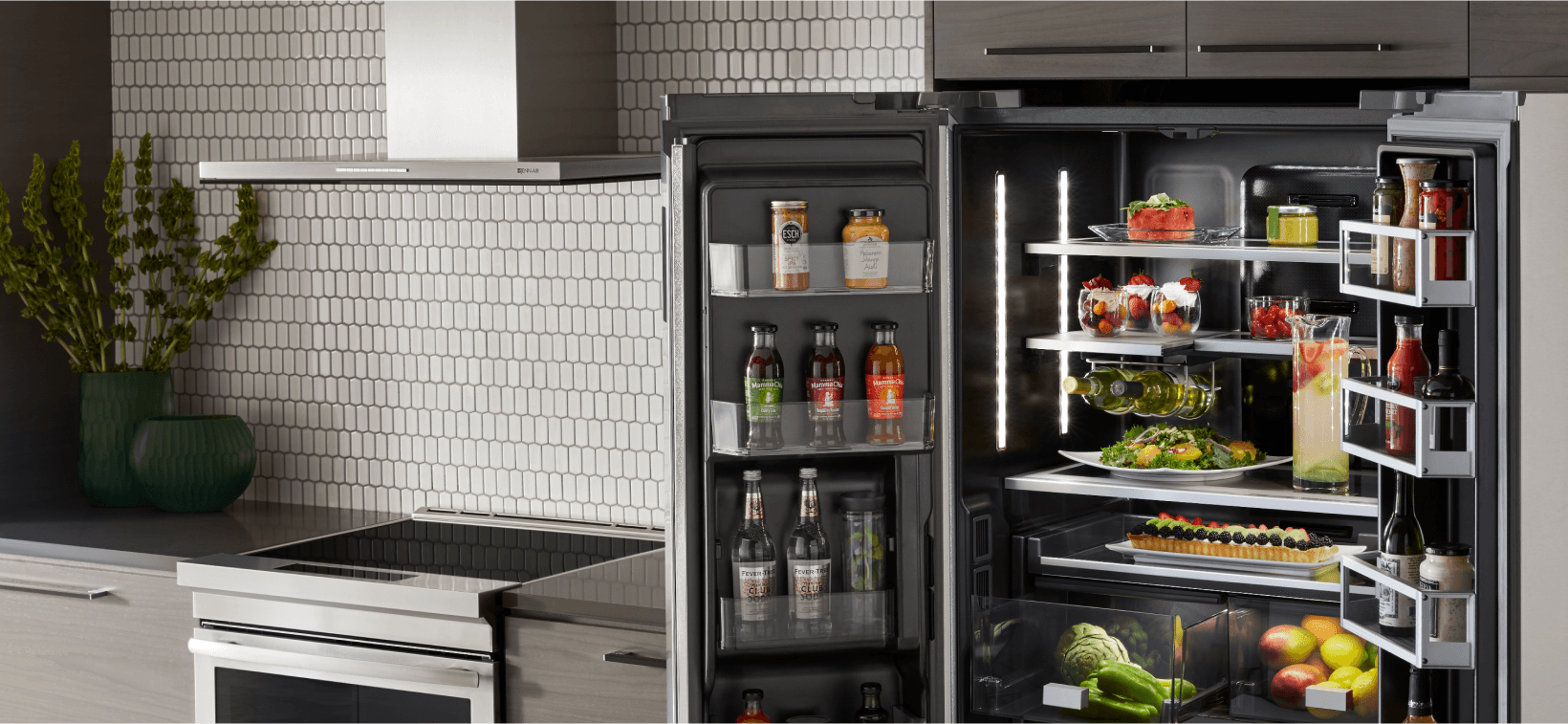  An opened JennAir refrigerator in a kitchen with a JennAir range. Inside the refrigerator are beverages, condiments, a flan, a plate of food, a pitcher of juice, mangoes and various produce, parfaits and cheeses.